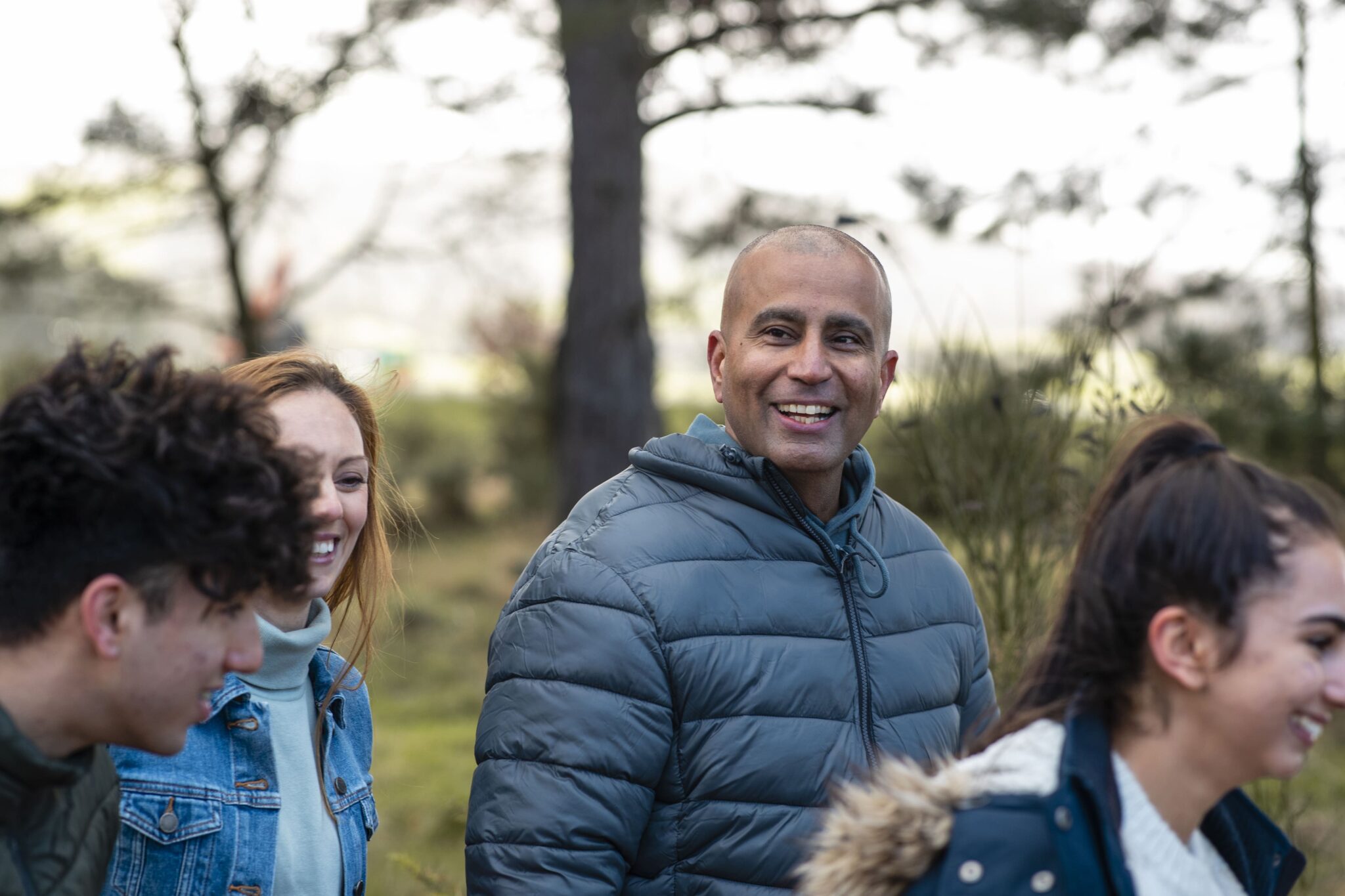 A group of people wearing jackets smiling and talking outdoors, with trees in the background.